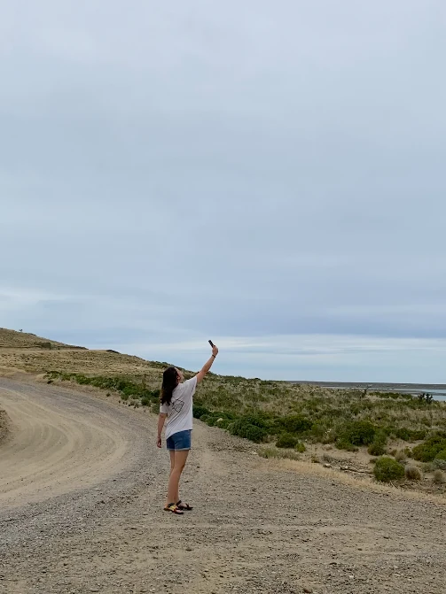 Viajera en remoto camino de ripio eleva el móvil buscando señal, con arbustos y el mar al fondo bajo un cielo gris