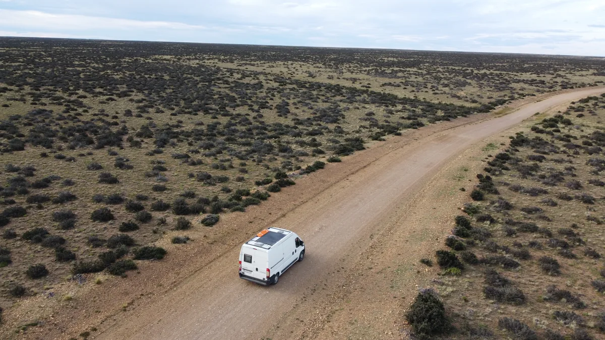 Furgoneta camper blanca con paneles solares rodando por pista de tierra en viaje overland por estepa árida