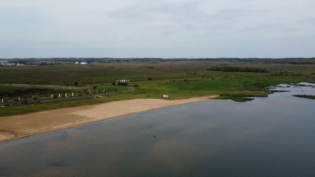 Furgoneta camper blanca en una playa de arena junto a un lago, rodeada de campos verdes y con edificios industriales al fondo