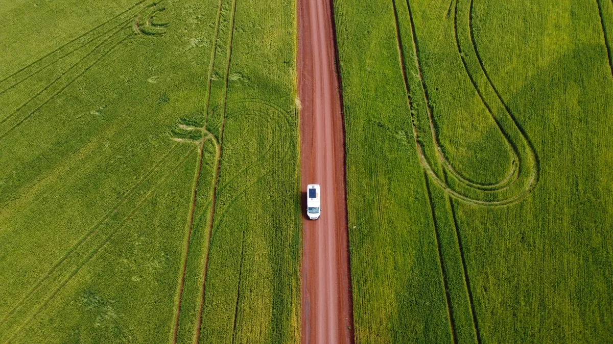 Vista aérea de una camper van blanca con panel solar rodando por un camino de tierra rojiza entre extensos campos verdes