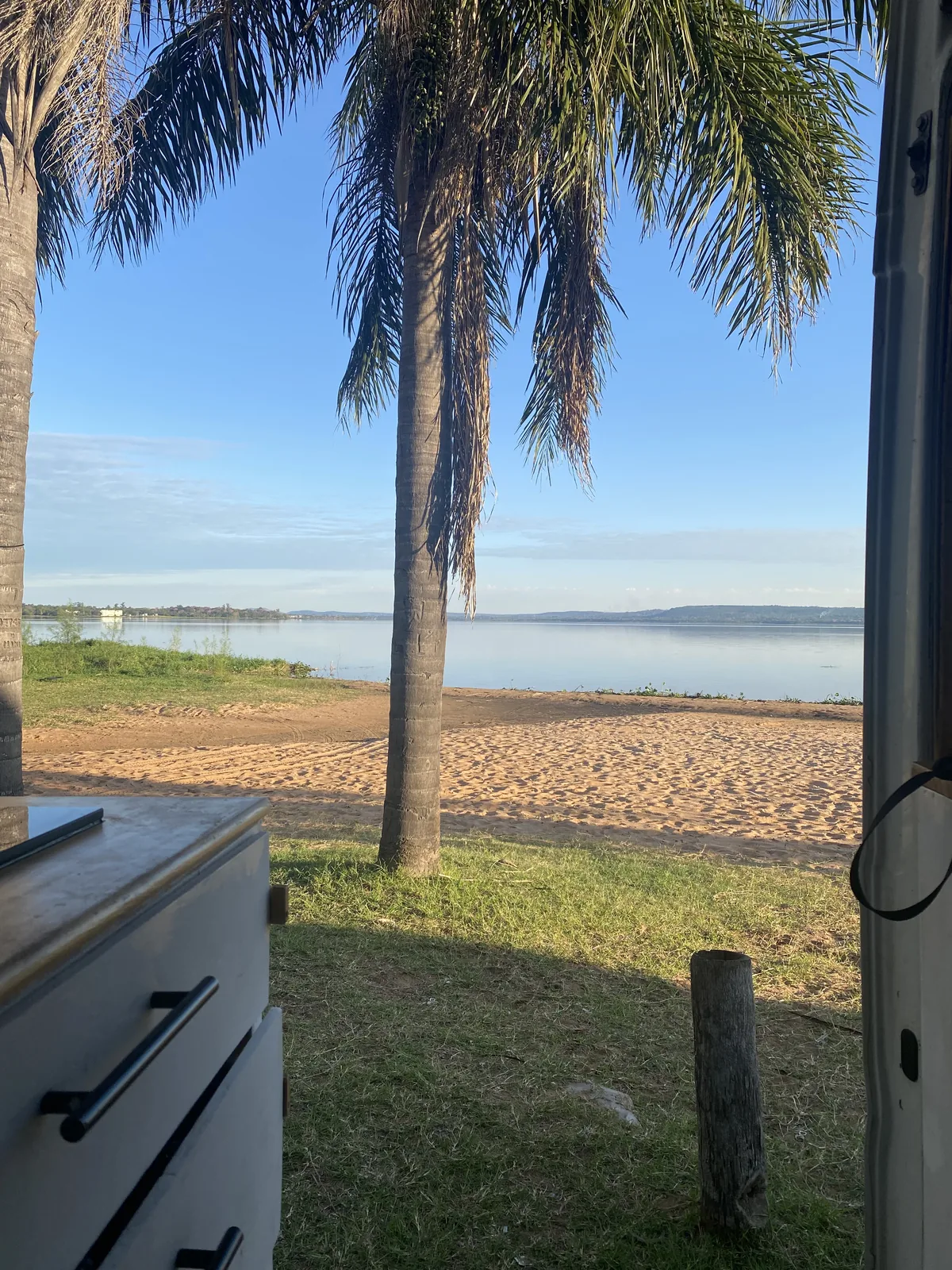 Vista desde la camper van: mueble con cajones, palmeras en la orilla, playa de arena y un gran lago azul