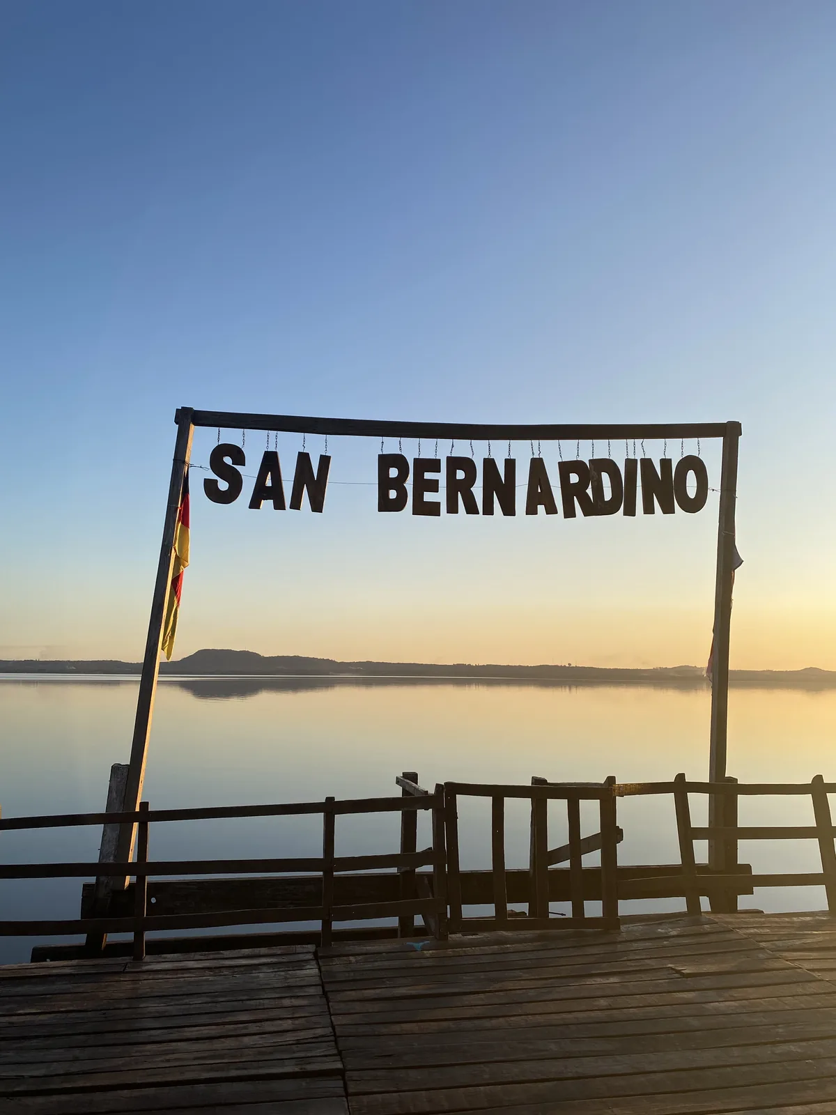 Muelle de madera con cartel de San Bernardino frente a un lago al amanecer, reflejando el cielo y el horizonte montañoso