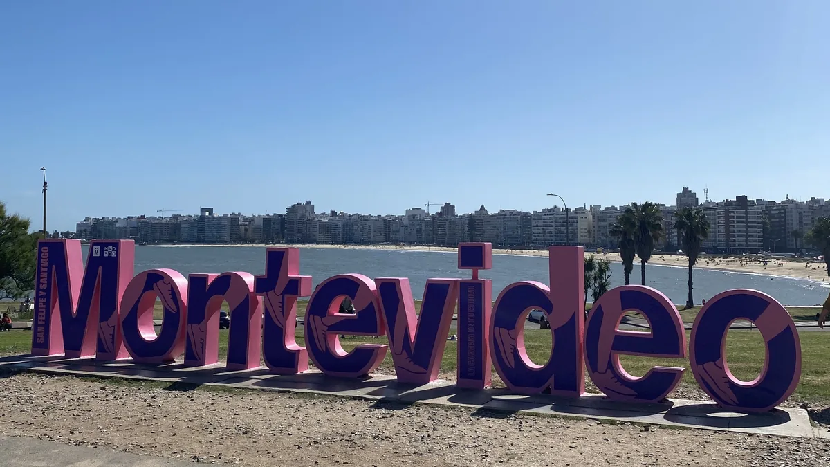 Icónicas letras de Montevideo en rosa y morado, con la rambla, playa y edificios de la capital uruguaya bajo un cielo azul