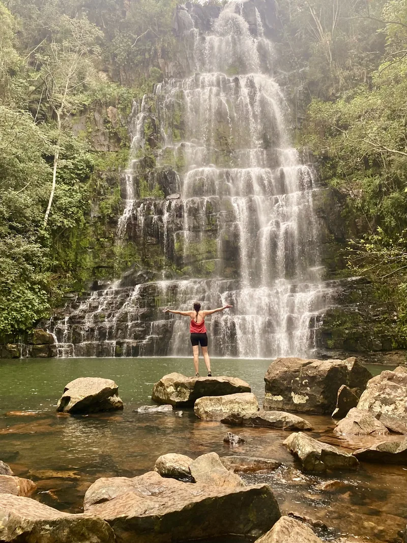 Mujer con brazos abiertos frente a una majestuosa cascada escalonada en la selva exuberante, con rocas en el agua