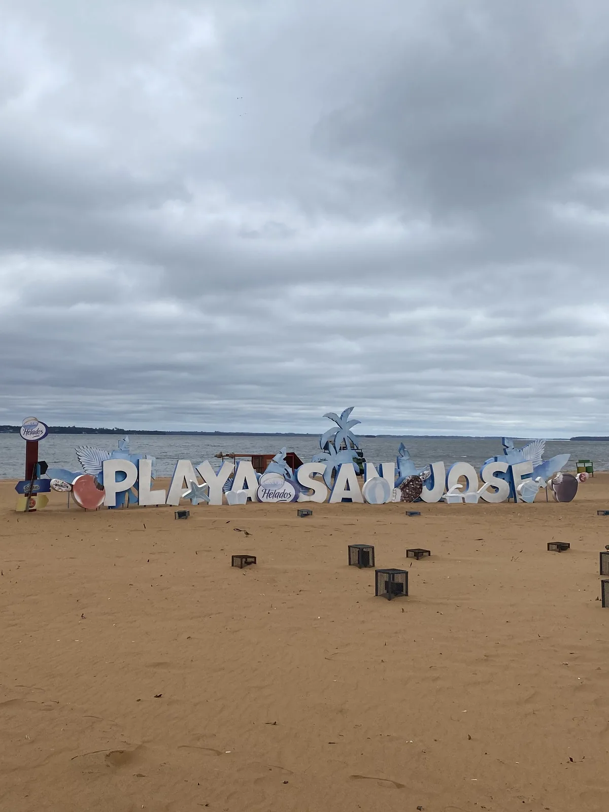 Letras blancas de "PLAYA SAN JOSE" con adornos en la arena de una playa fluvial, bajo un cielo gris y cubierto.