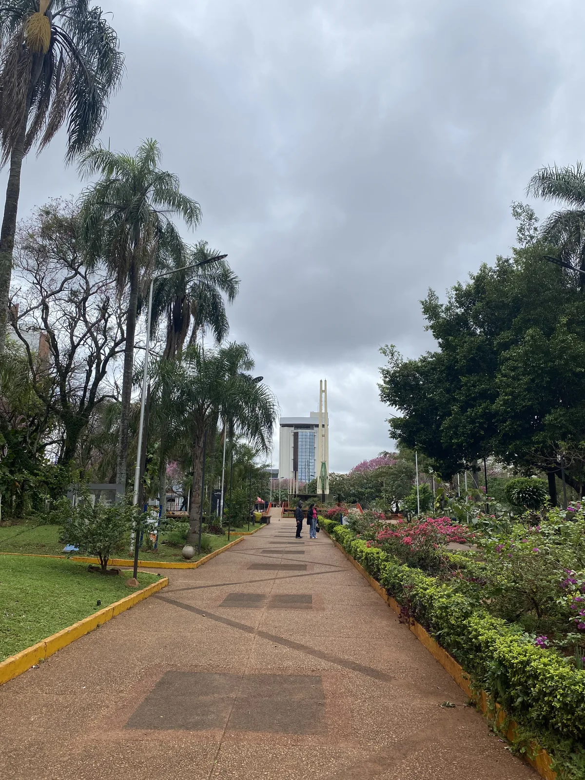 Paseo en parque de Asunción, Paraguay, con palmeras, flores coloridas y un edificio moderno bajo un cielo gris