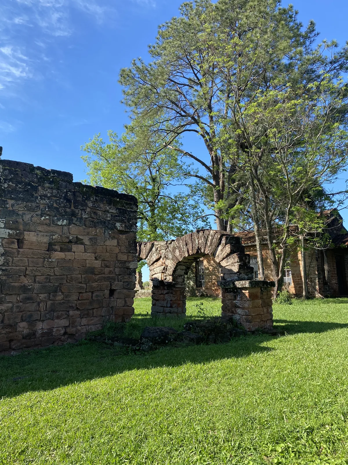 Ruinas de antiguas estructuras de piedra rojiza con un arco y muro, rodeadas de césped verde y grandes árboles