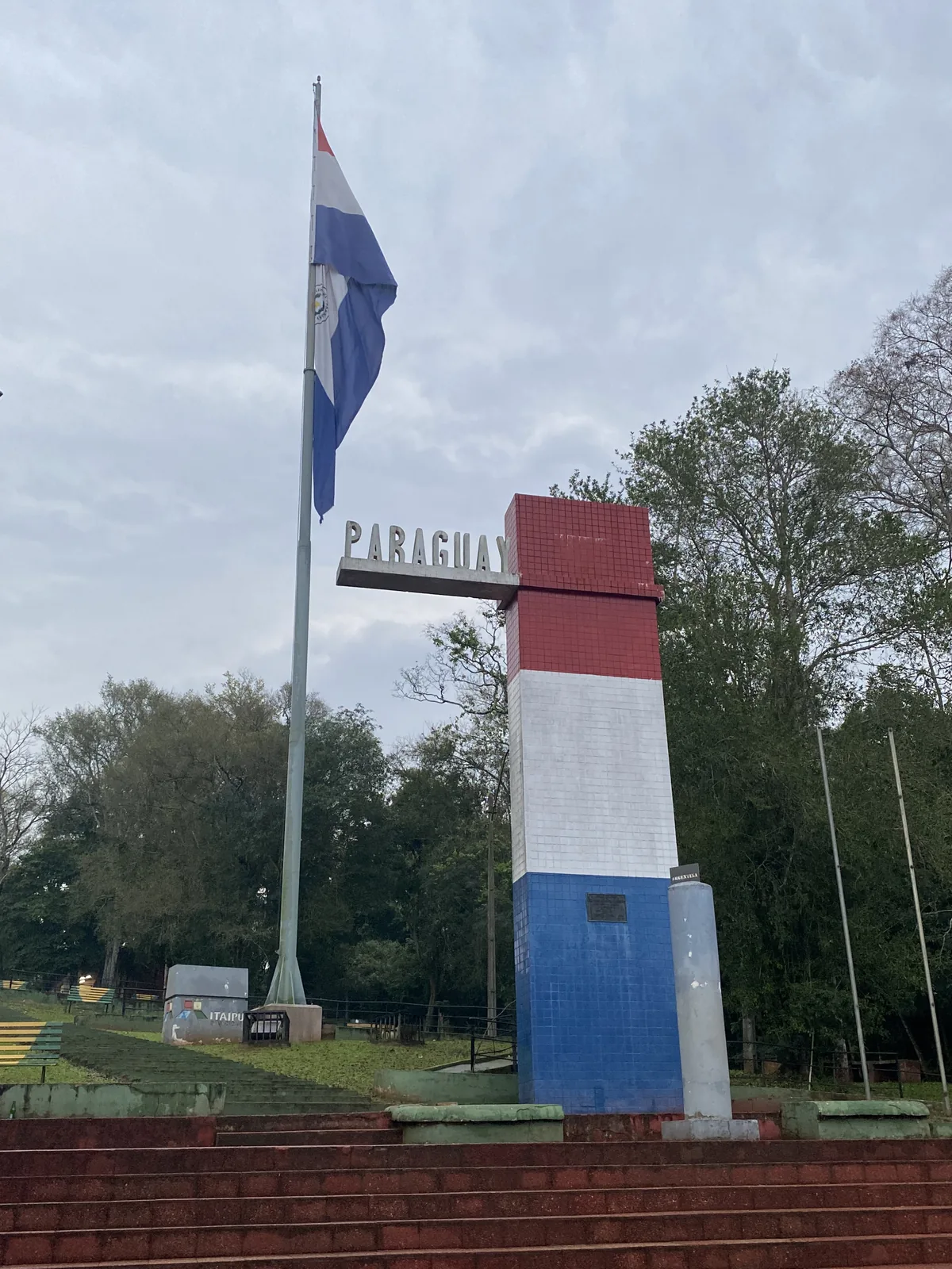 Hito de Paraguay con bandera nacional y monumento tricolor cerca de Itaipu, visto desde escaleras en un día nublado