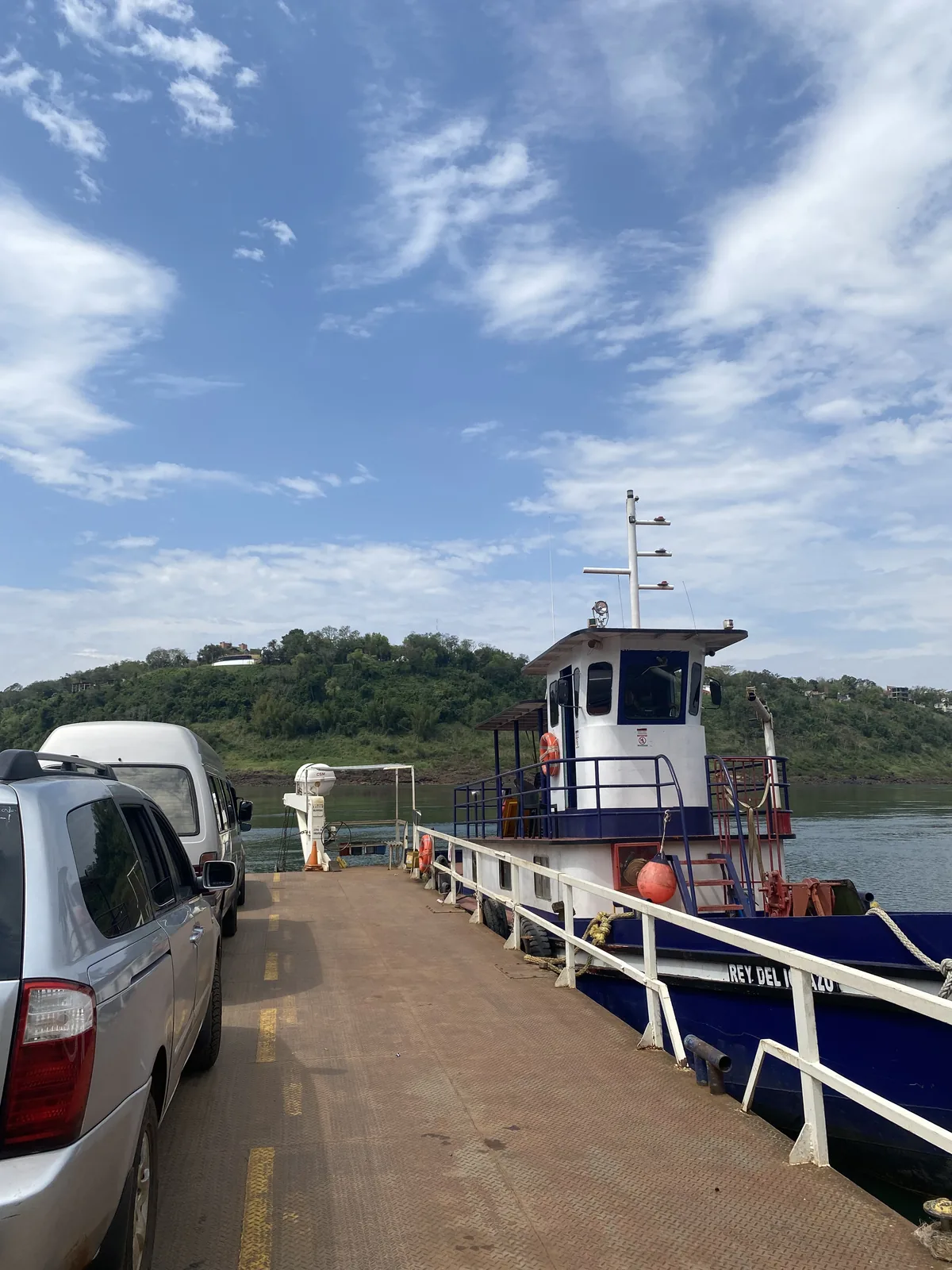 Minivan plata y furgoneta overland con baca en el ferry Rey del Azu, cruzando un río ancho entre colinas verdes