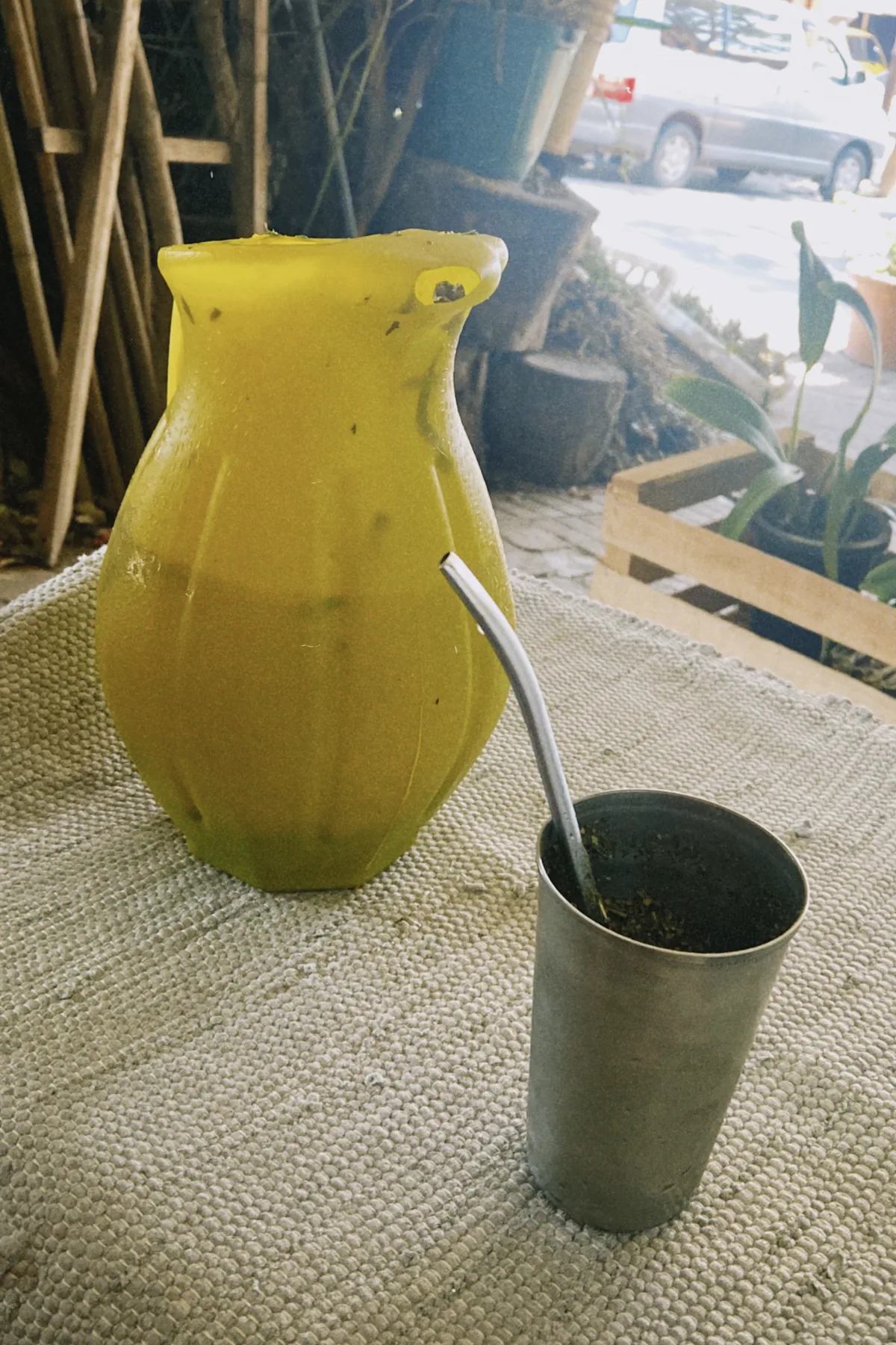 Jarra amarilla de tereré con bombilla y mate de metal sobre una mesa, con plantas y un coche en el fondo de un patio