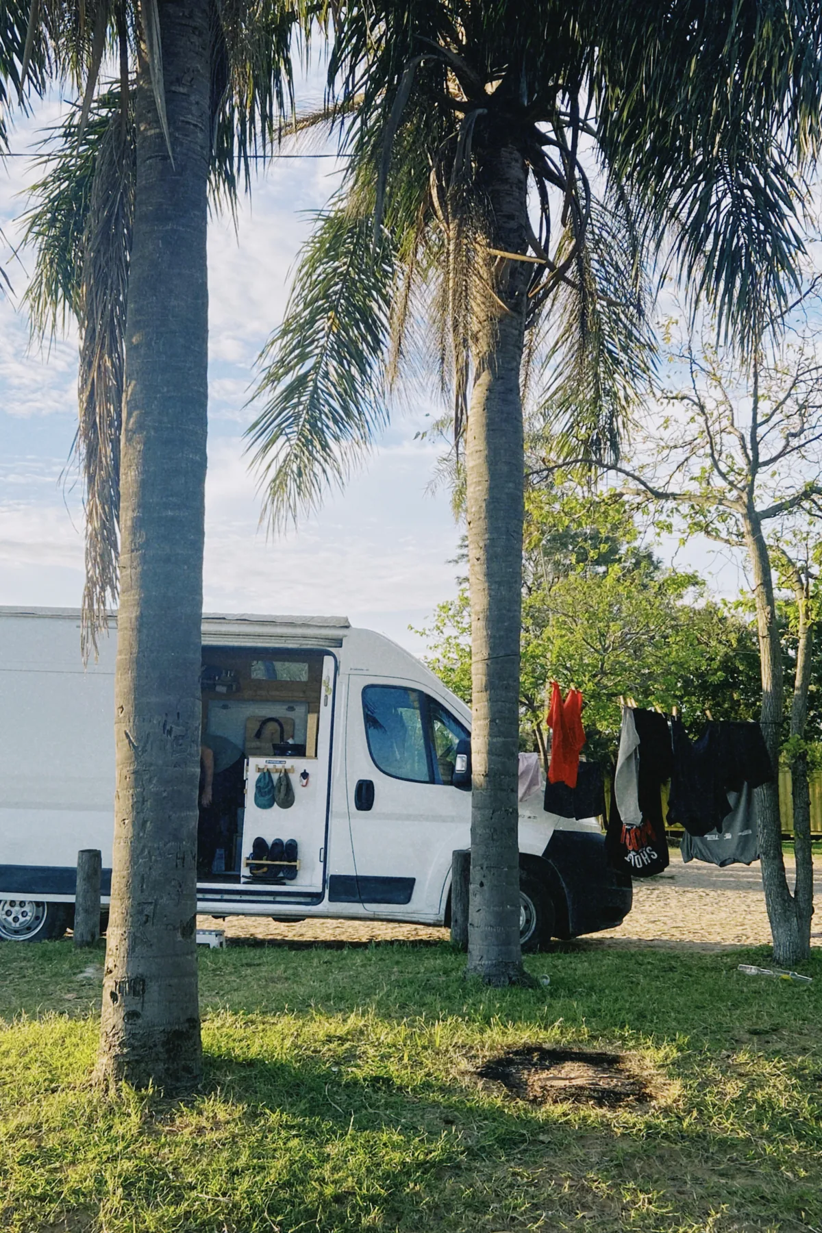 Van camper blanca estacionada en un entorno tropical con palmeras. Puerta lateral abierta mostrando interior y sandalias