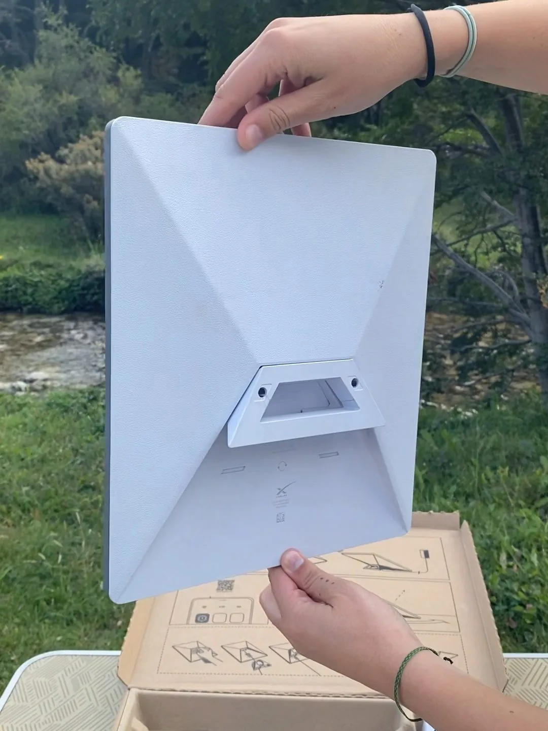 Manos mostrando la antena Starlink portátil (modelo mini) en un campamento junto a un río, perfecta para internet en ruta