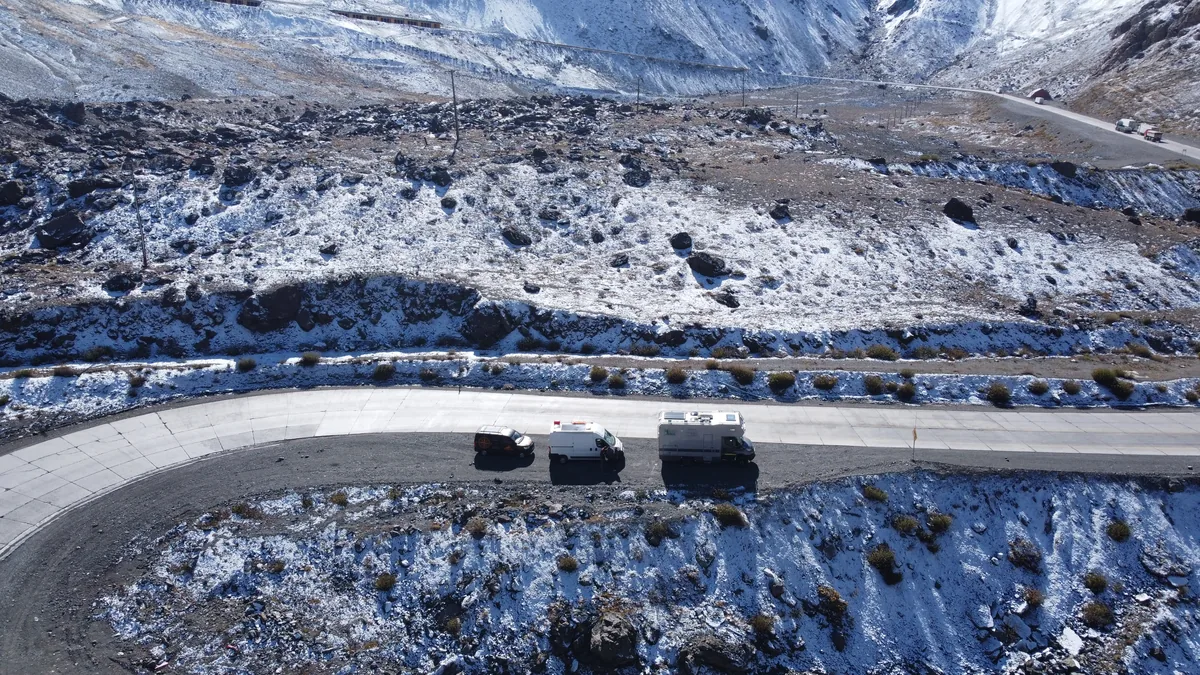 Vista aérea de tres vehículos overland, una furgoneta camper y un camión expedicionario, en carretera andina nevada