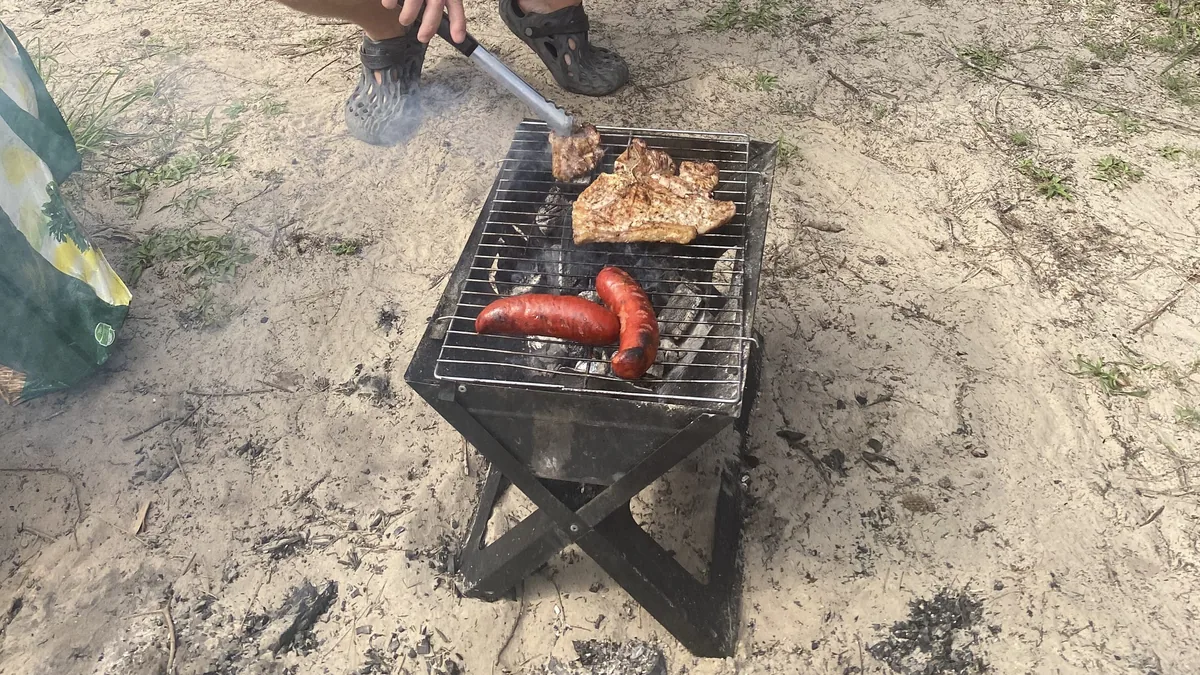Cocina en ruta: persona preparando un asado con chorizos y chuletas en una parrilla portátil sobre arena