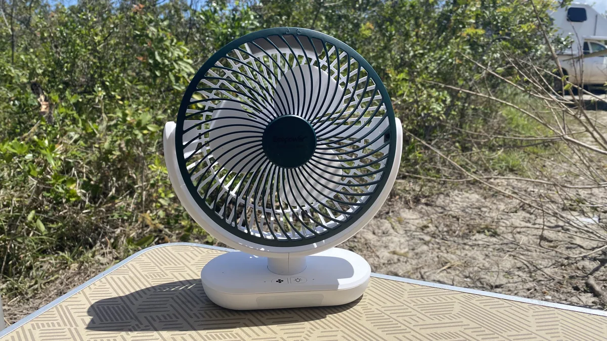 Ventilador recargable blanco y verde sobre mesa de camping, con camioneta camper blanca en un entorno natural