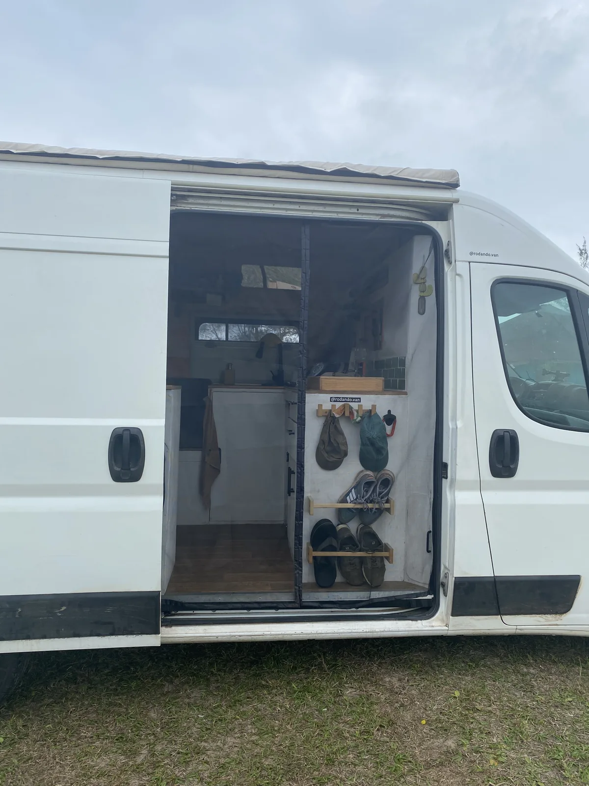 Furgoneta camper blanca con puerta corredera abierta. Interior con cocina, mosquitera, y estante de madera para zapatos