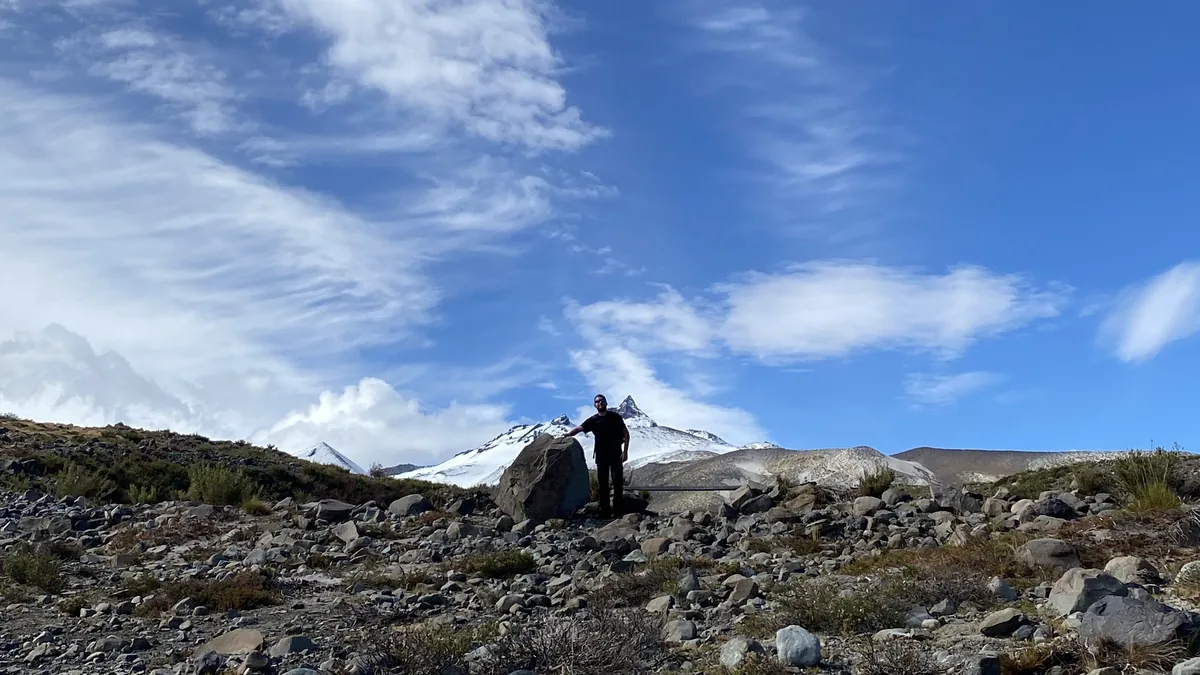 Explorador overland junto a roca en terreno rocoso, con volcán nevado y montañas de fondo bajo un cielo azul vibrante