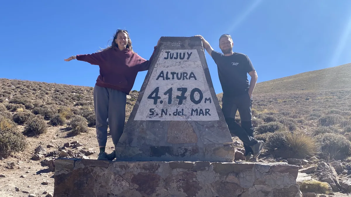 Viajeros overland posando en el mojón de altura 4170m en Jujuy, Argentina, entre montañas áridas bajo cielo azul