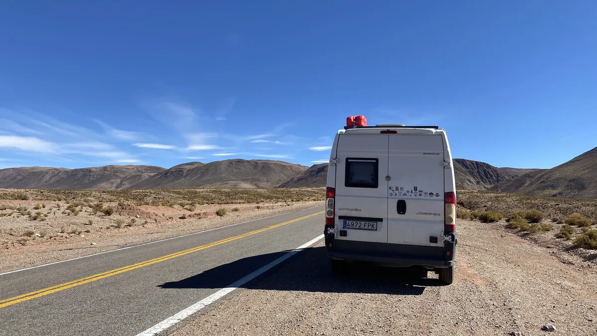 Furgoneta camper Citroën Jumper blanca con bidones en el techo, en ruta por desierto montañoso bajo cielo azul