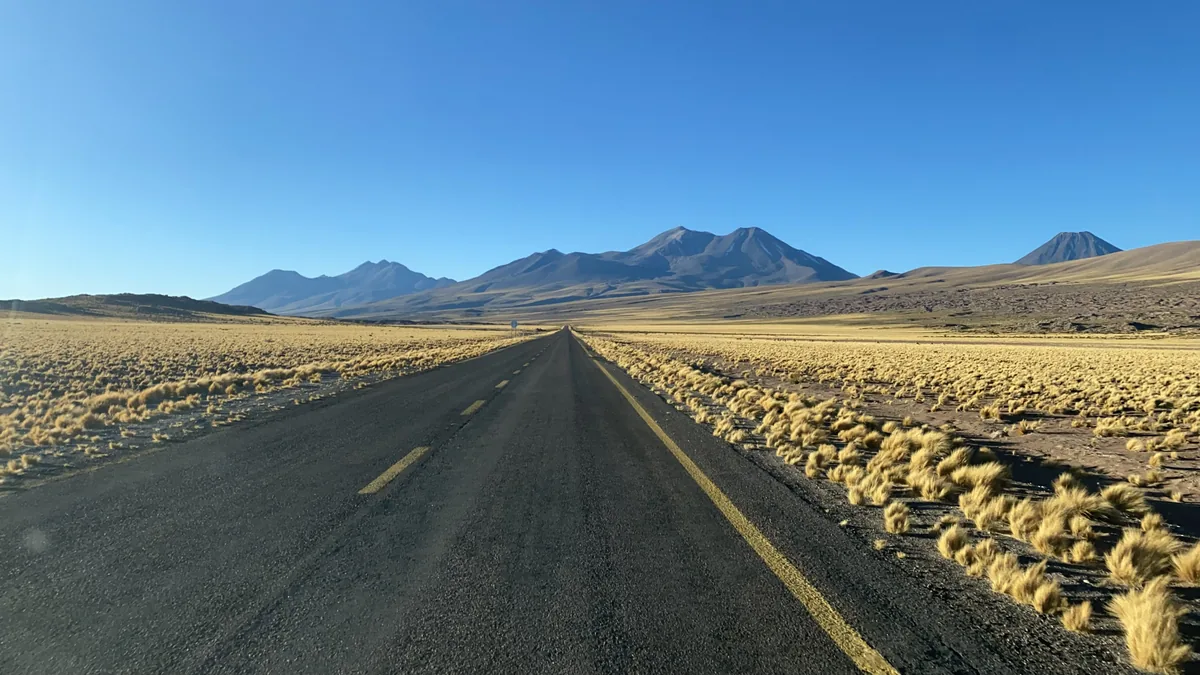 Carretera en el altiplano andino, rodeada de pastos dorados y volcanes bajo un cielo azul intenso