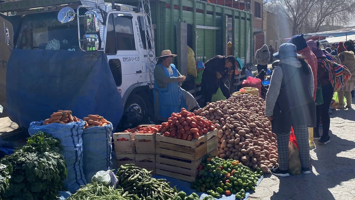Animado mercado andino con un camión japonés, vendedora ofreciendo montañas de papas, tomates, zanahorias y verduras frescas