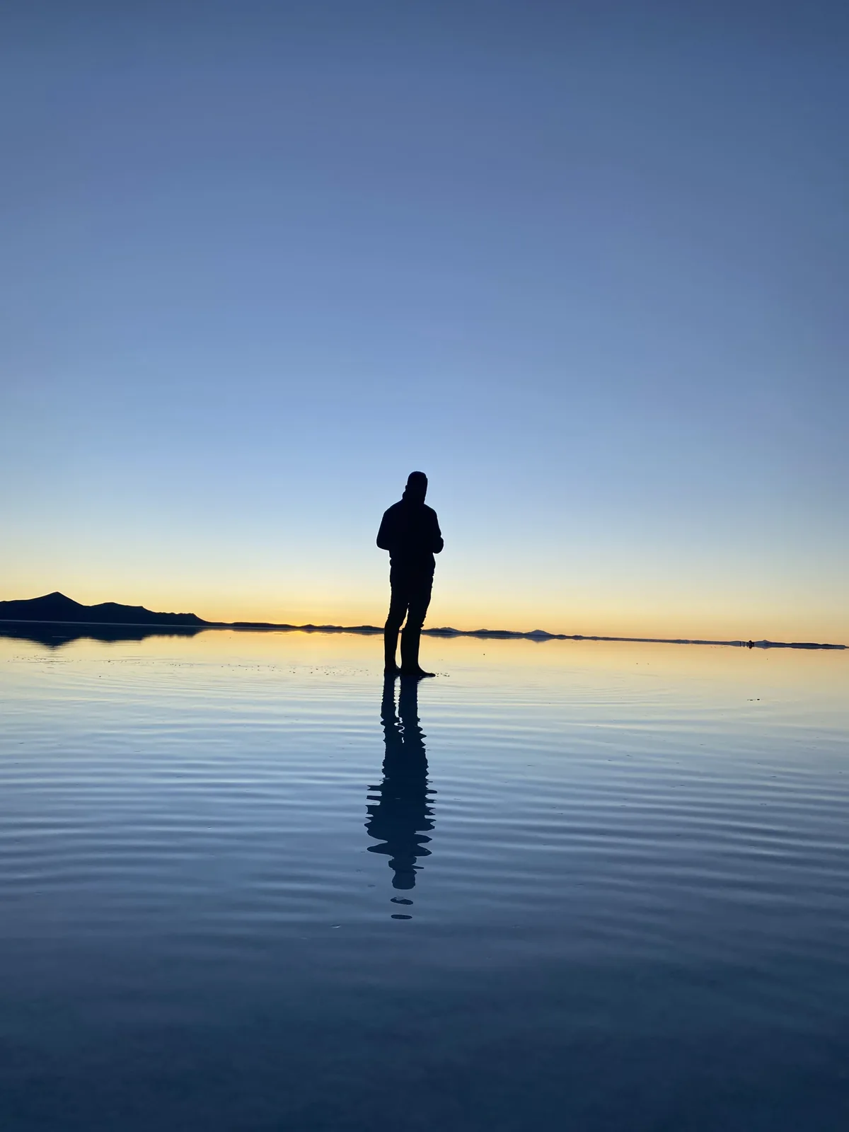 Silueta de viajero con botas en el Salar de Uyuni al amanecer, reflejo perfecto en el agua, montañas lejanas