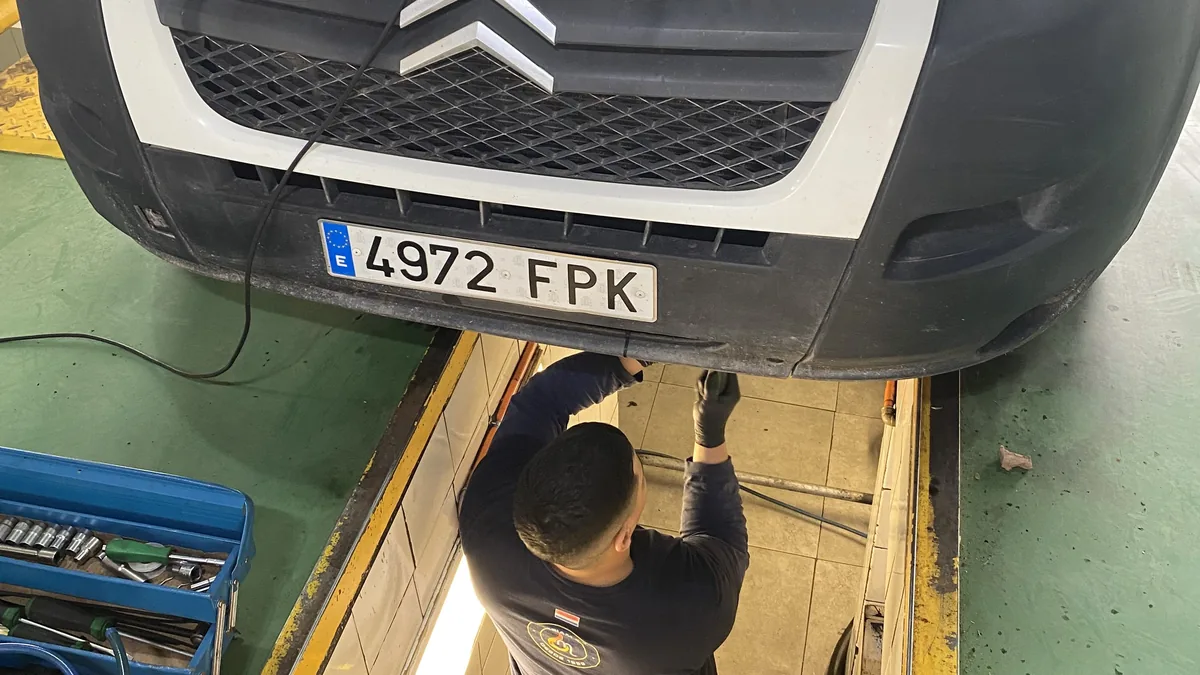 Mecánico con guantes revisa conexión eléctrica bajo el parachoques de una furgoneta Citroën camper blanca en taller