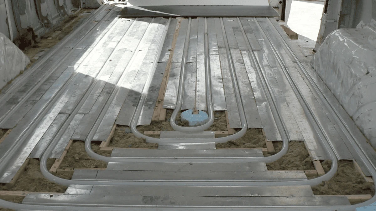 Detalle del suelo de una furgoneta camper en construcción, mostrando la instalación de calefacción radiante con tuberías