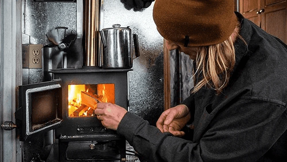 Overlander añadiendo leña a una estufa de leña dentro de su camper van, con una tetera calentándose