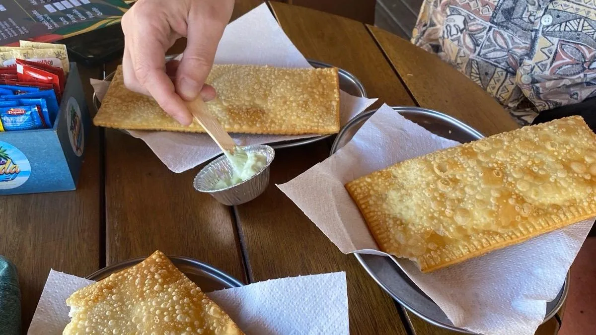 Disfrutando tres pastéis fritos dorados en una mesa de madera. Una mano sumerge el pastel en salsa verde. Sabor local