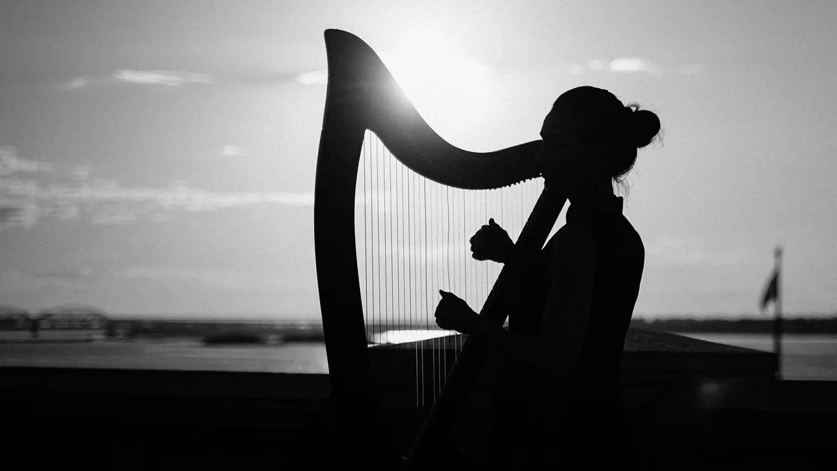 Silueta de mujer tocando el arpa al atardecer, el sol brillando a través de sus cuerdas y un río al fondo