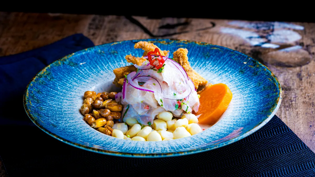 Ceviche de pescado fresco con cebolla morada, camote, choclo, cancha y chicharrón, en un plato azul rústico peruano