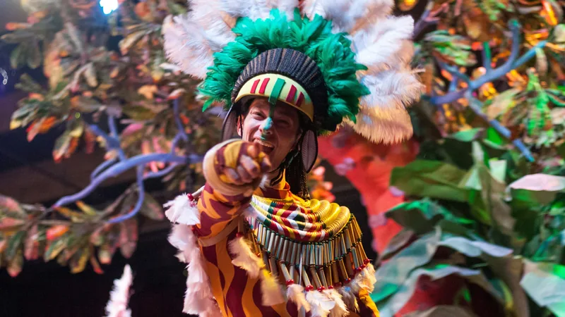 Sonriente personaje indígena con tocado de plumas verdes y blancas, traje ceremonial, apuntando en un festival de selva