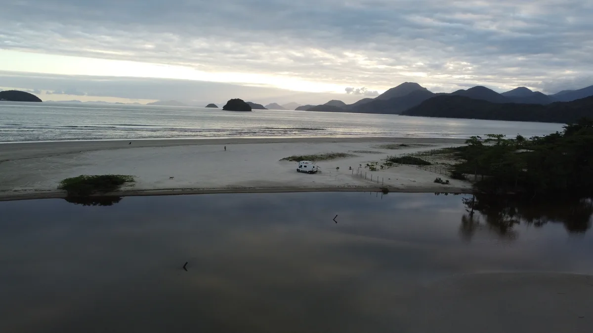 Campervan blanca estacionada en playa desierta junto a una laguna, con el mar, islas y montañas en un amanecer nublado