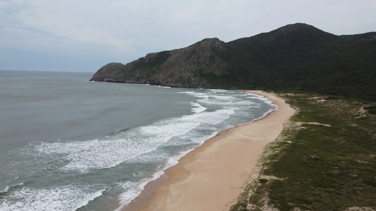 Vista aérea de una extensa playa de arena clara y olas en un día nublado, flanqueada por grandes montañas cubiertas de densa