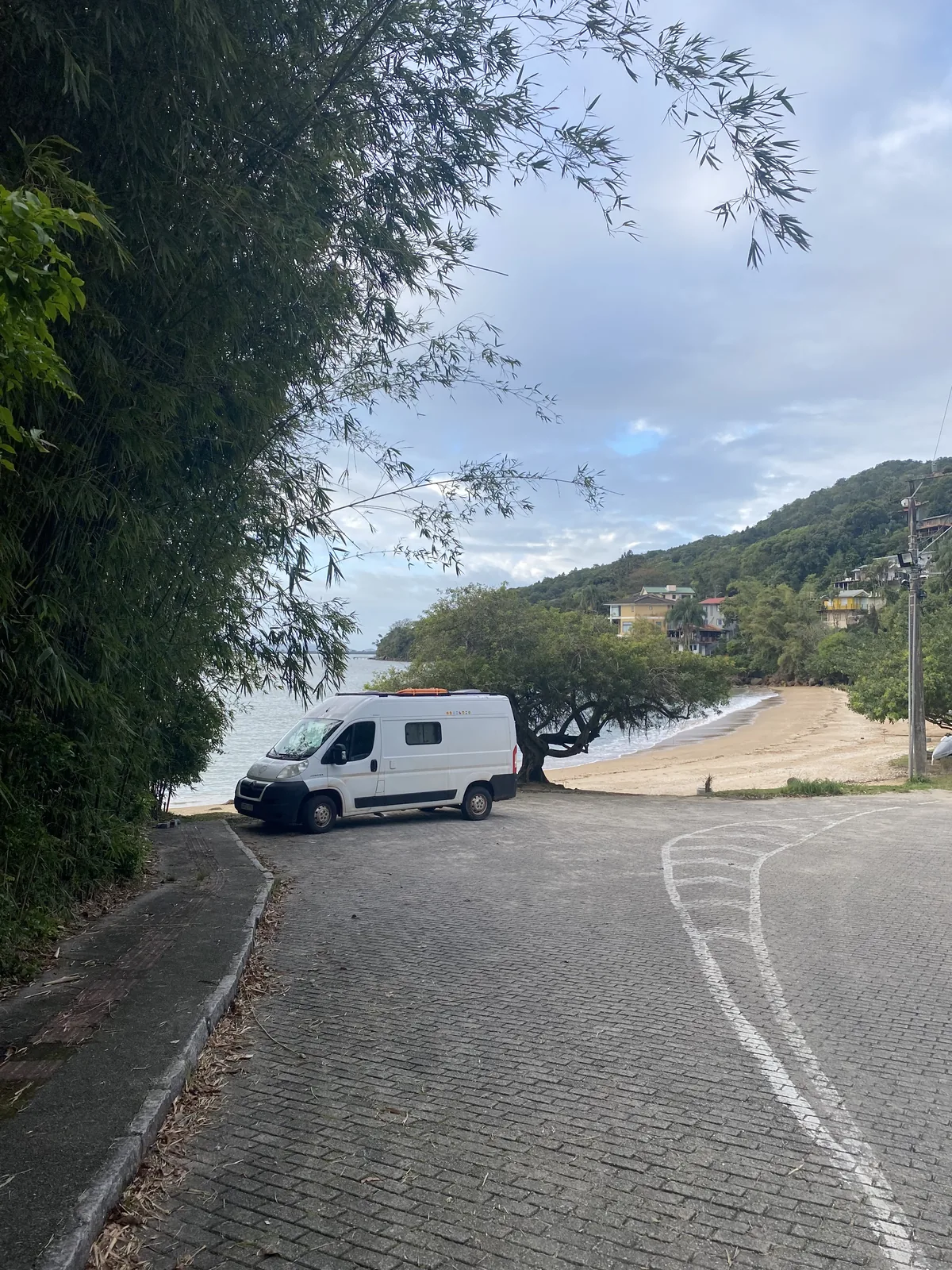 Furgoneta camper blanca con baca en una playa tropical de arena y bambú, con casas en las colinas y mar tranquilo