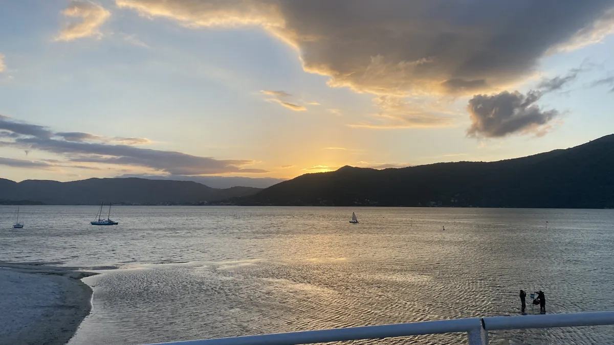 Panorama al atardecer de una bahía serena con veleros, montañas y siluetas de personas explorando la orilla. Viaje overland