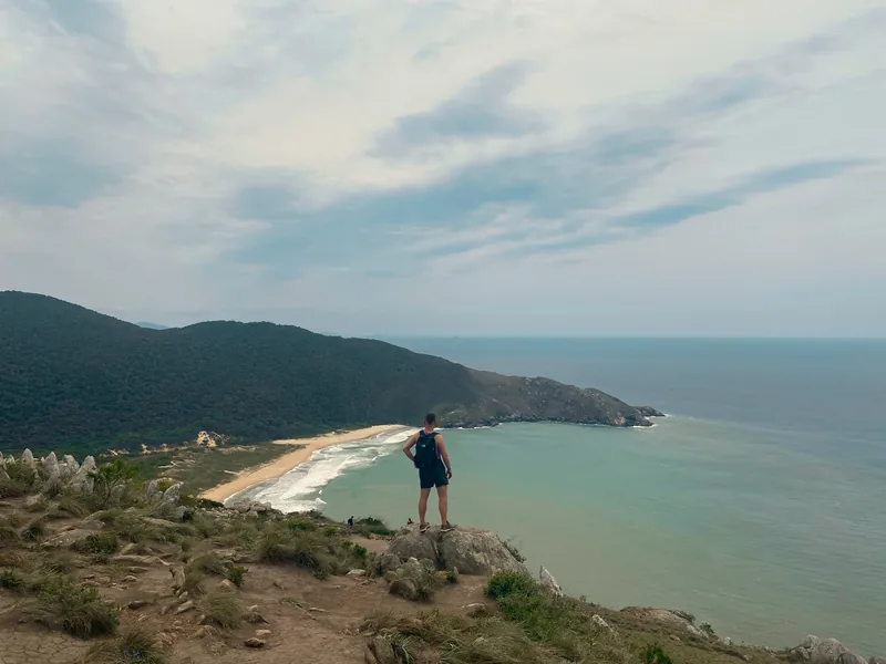 Viajero en un mirador rocoso con mochila, admirando una playa de arena dorada y el mar turquesa rodeado de colinas verdes
