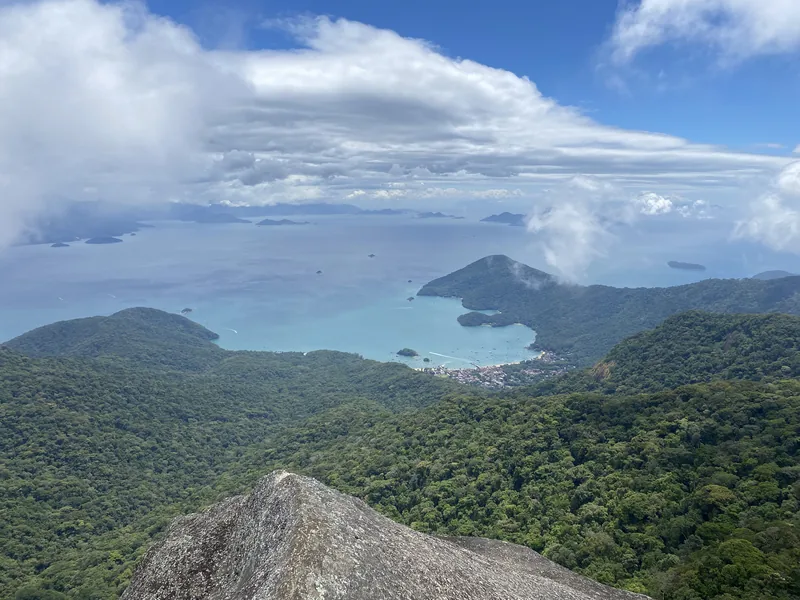Vista desde cumbre rocosa de bahía tropical con pueblo costero, densa selva, islas y cielo parcialmente cubierto