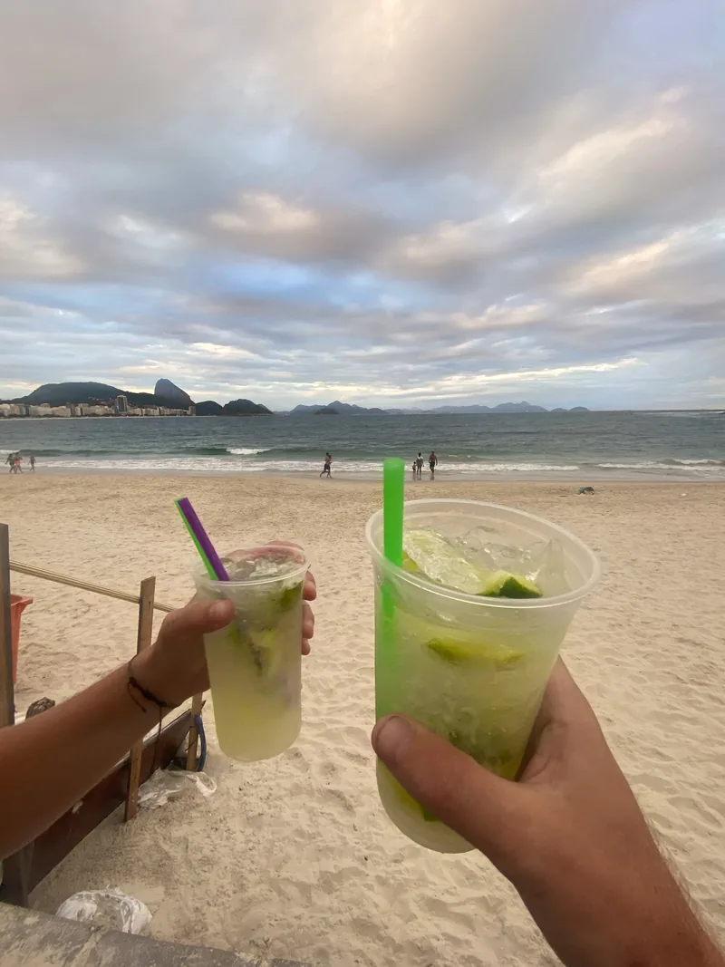 Manos brindando con caipirinhas en Copacabana, Río de Janeiro, frente al Pan de Azúcar y el mar bajo un cielo nublado