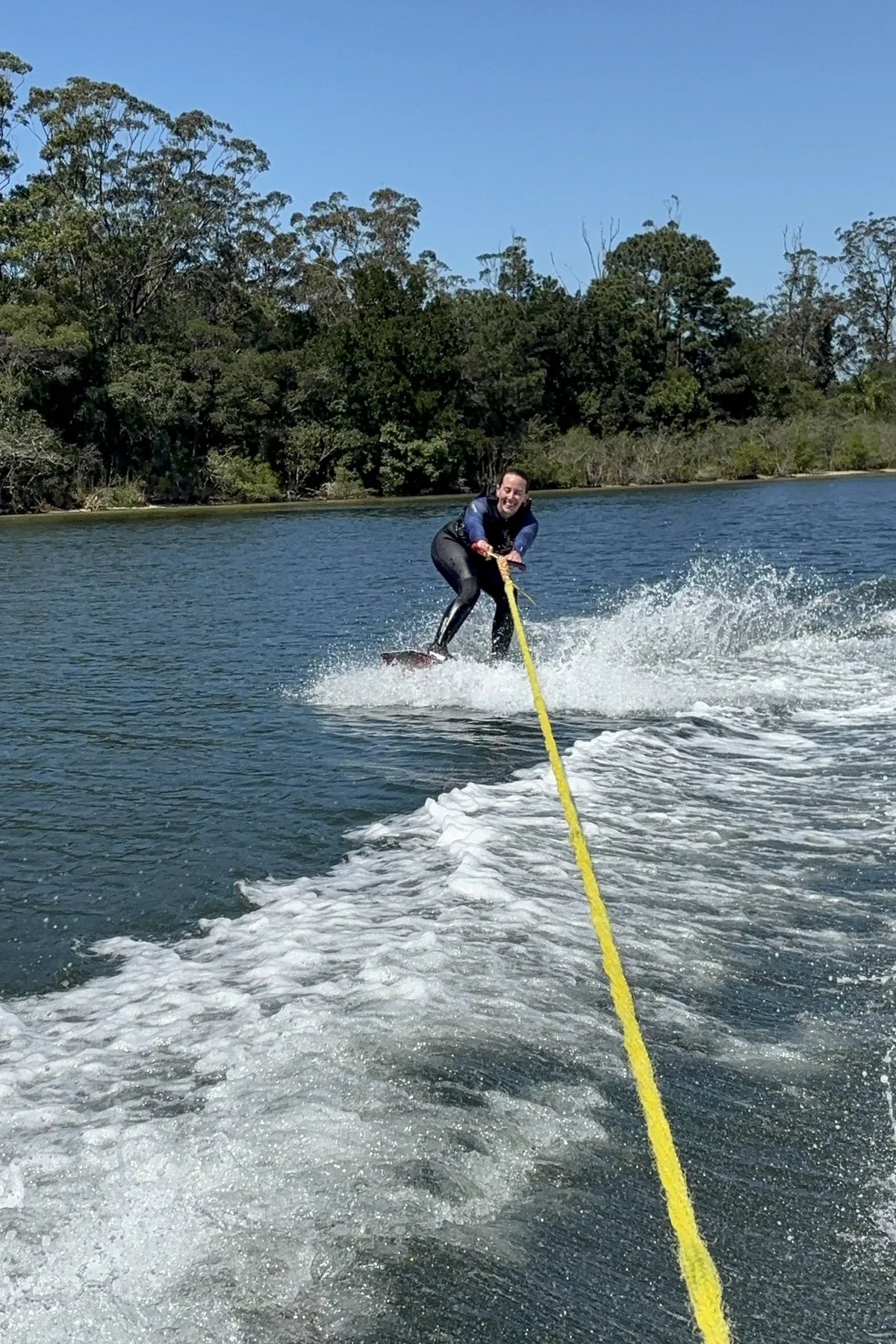 Mujer sonriente en traje de neopreno wakeboarding en río, con bosque frondoso y cielo azul claro de fondo