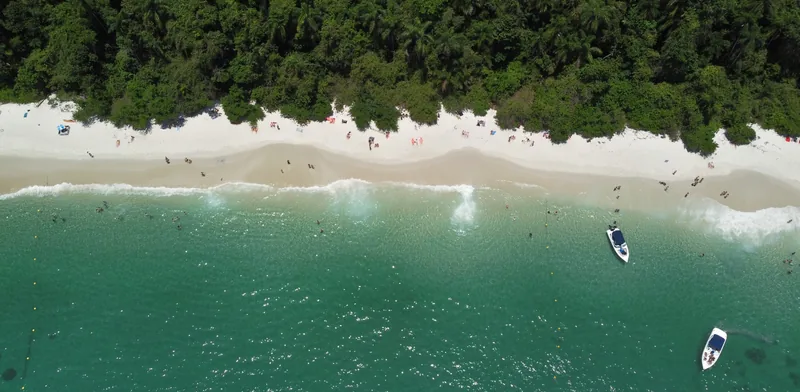 Vista aérea de playa de arena blanca, vegetación densa, agua turquesa, gente y dos barcos