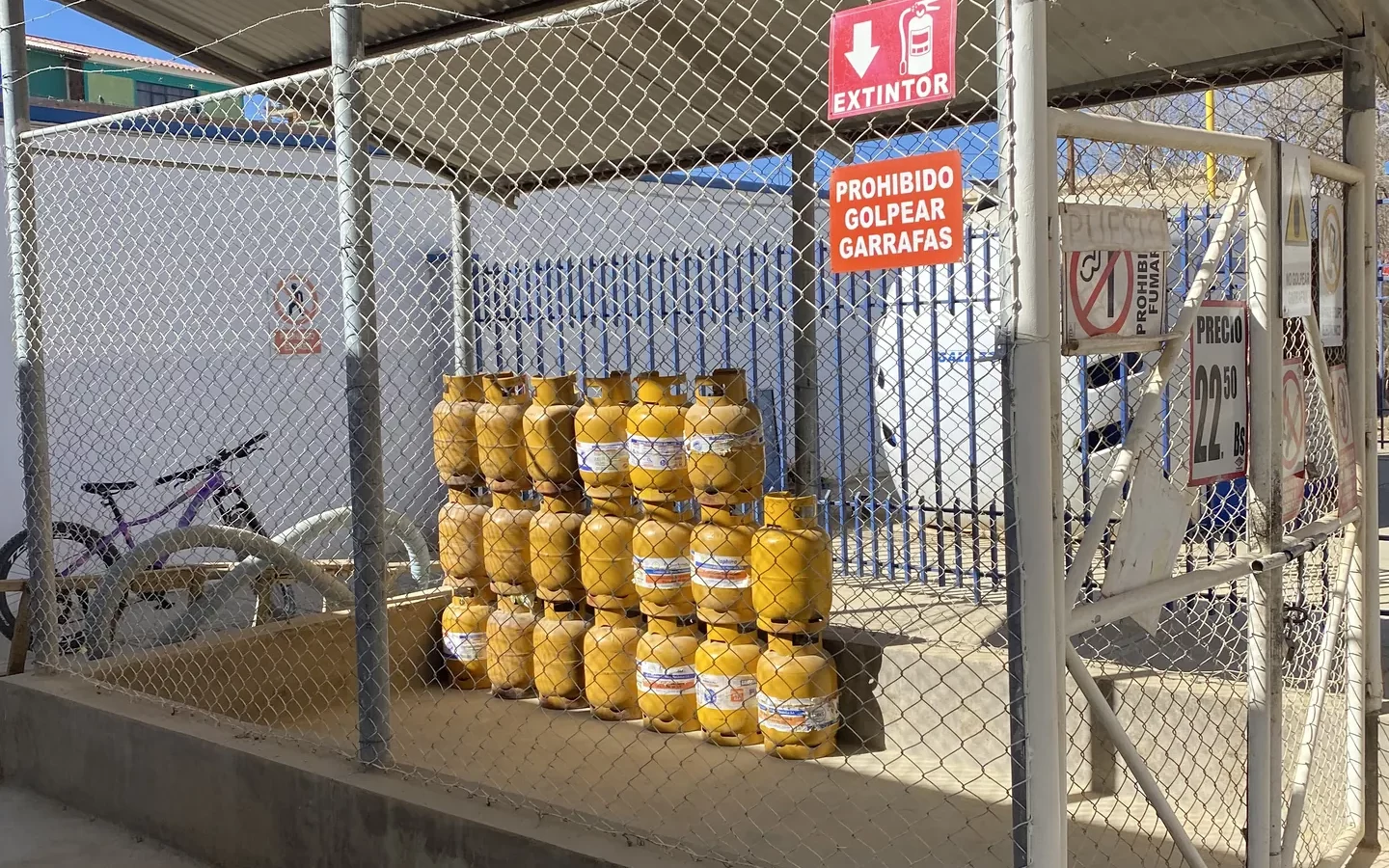Almacén vallado con garrafas de gas amarillas apiladas y dos carteles de seguridad