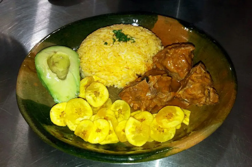 Seco de chivo servido con arroz, aguacate y plátano frito