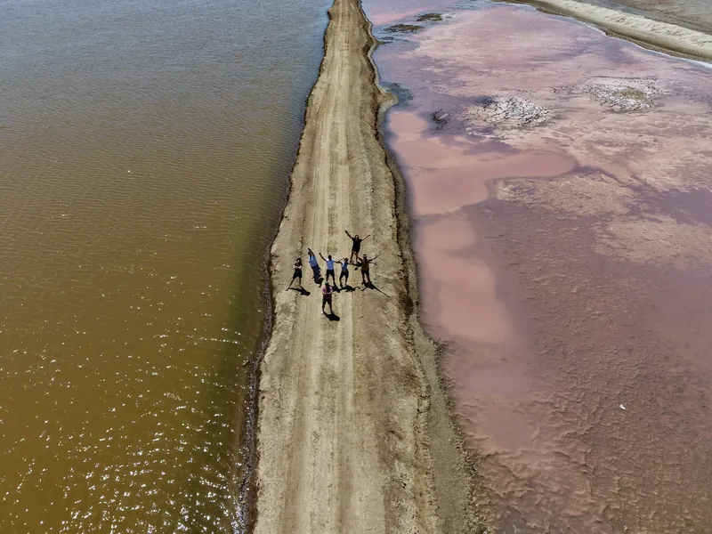 Vista aérea de un grupo de personas con los brazos abiertos sobre un camino entre una laguna marrón y unas salinas rosas en la costa de Ica, Perú