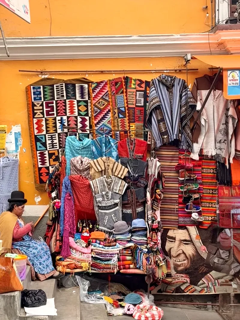 Puesto de ponchos, mantas y gorros de lana junto a un mural en una calle de La Paz, Bolivia